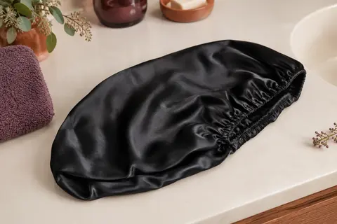 Bonnet de nuit long en satin noir posé dans une salle de bain lumineuse aux tons ivoire et prune.